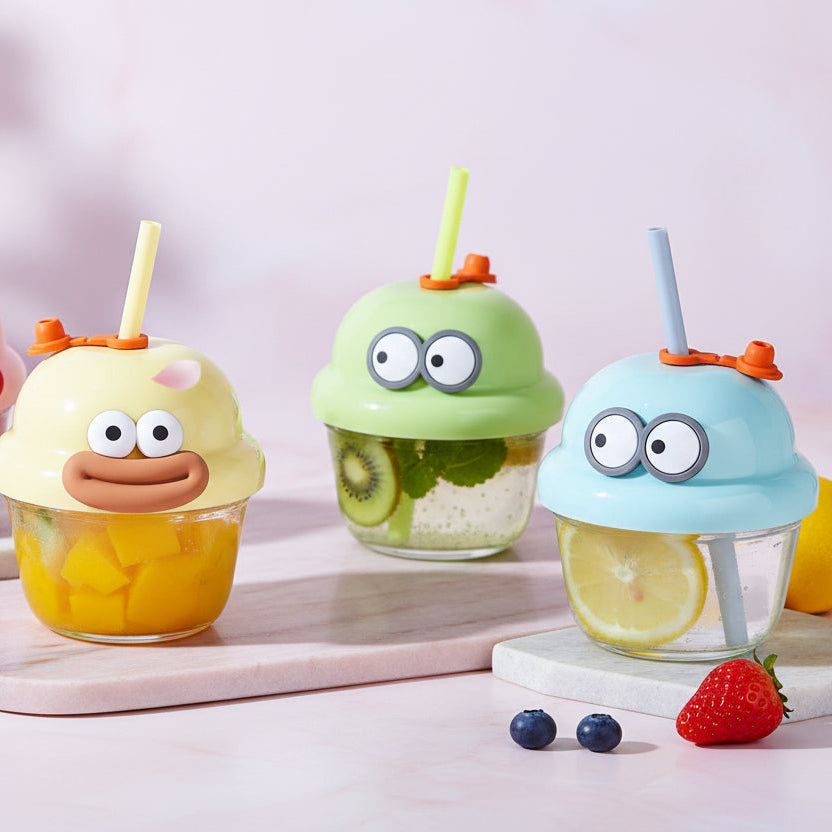 Four cartoon ice cream cup drink holders in pink, yellow, green, and blue, each with a matching straw and small cup underneath, arranged on a tabletop with books in the background. Tumbler