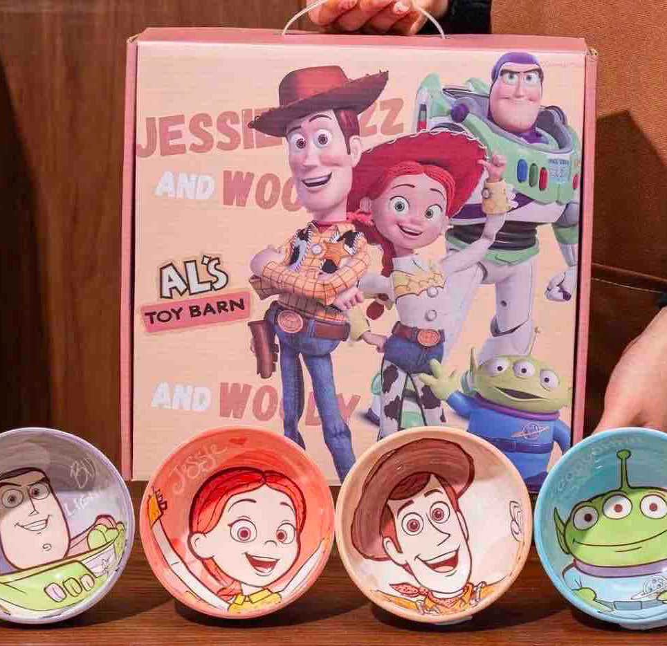 A Toy Barn gift set featuring a pink box with Buzz Lightyear, Jessie, Woody, and Alien characters, with four small illustrated bowls placed in front on a wooden surface.