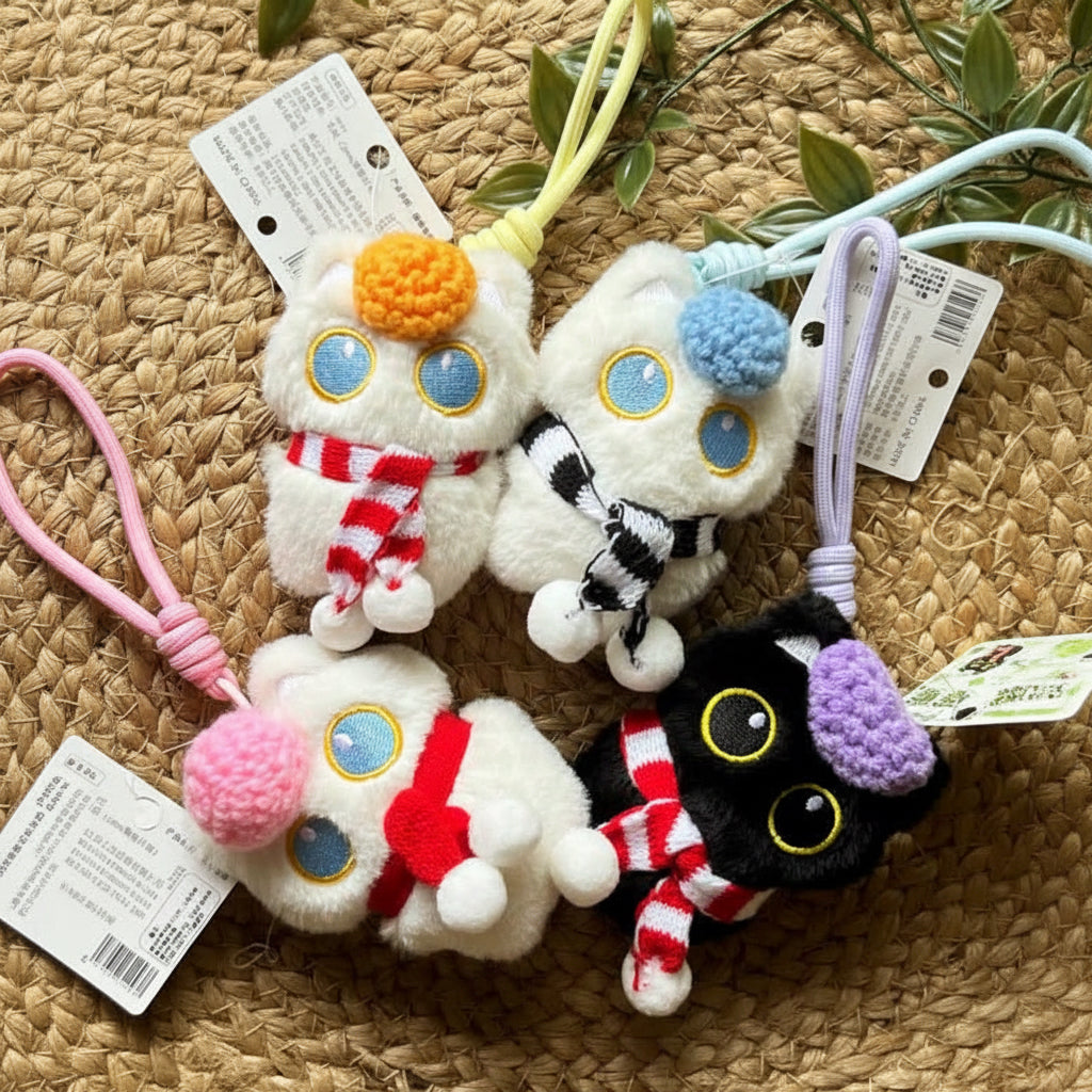 Four small plush character keychains or bag charms arranged on a woven straw surface with green leaves around them. The set includes two white characters with blue eyes and red-and-white scarves, one white character with a black-and-white scarf, and one black character with a purple bow and red-and-white scarf. Gift items